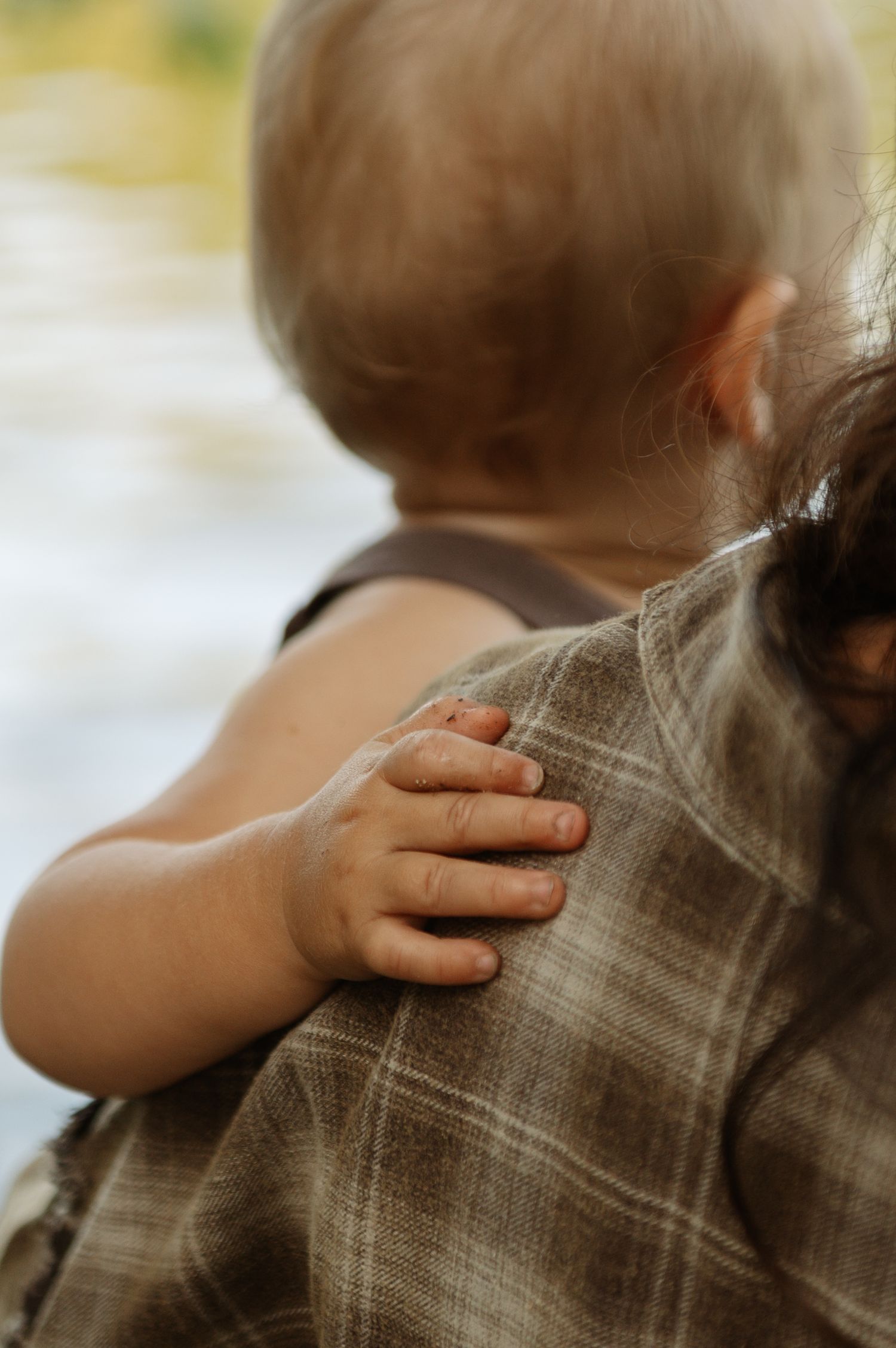 A baby rests its tiny hand on an adult's plaid shirt shoulder while being held.