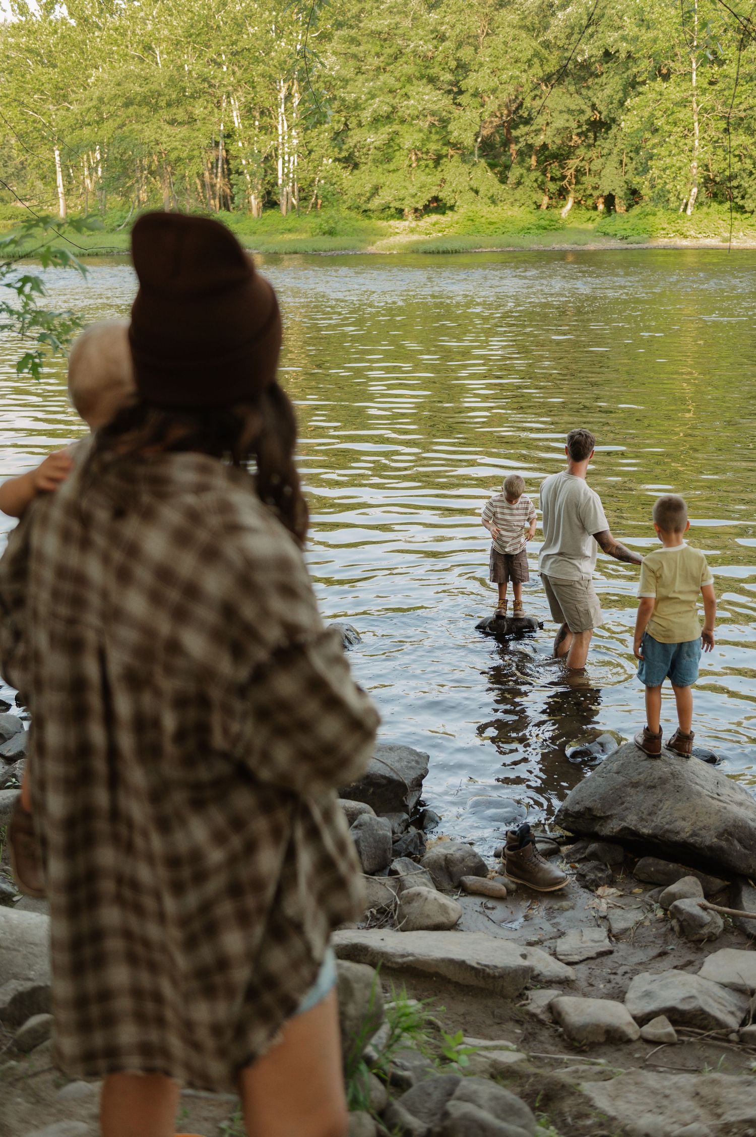 Family wading in a calm river surrounded by lush green trees, with a woman in plaid shirt watching from rocky shore.