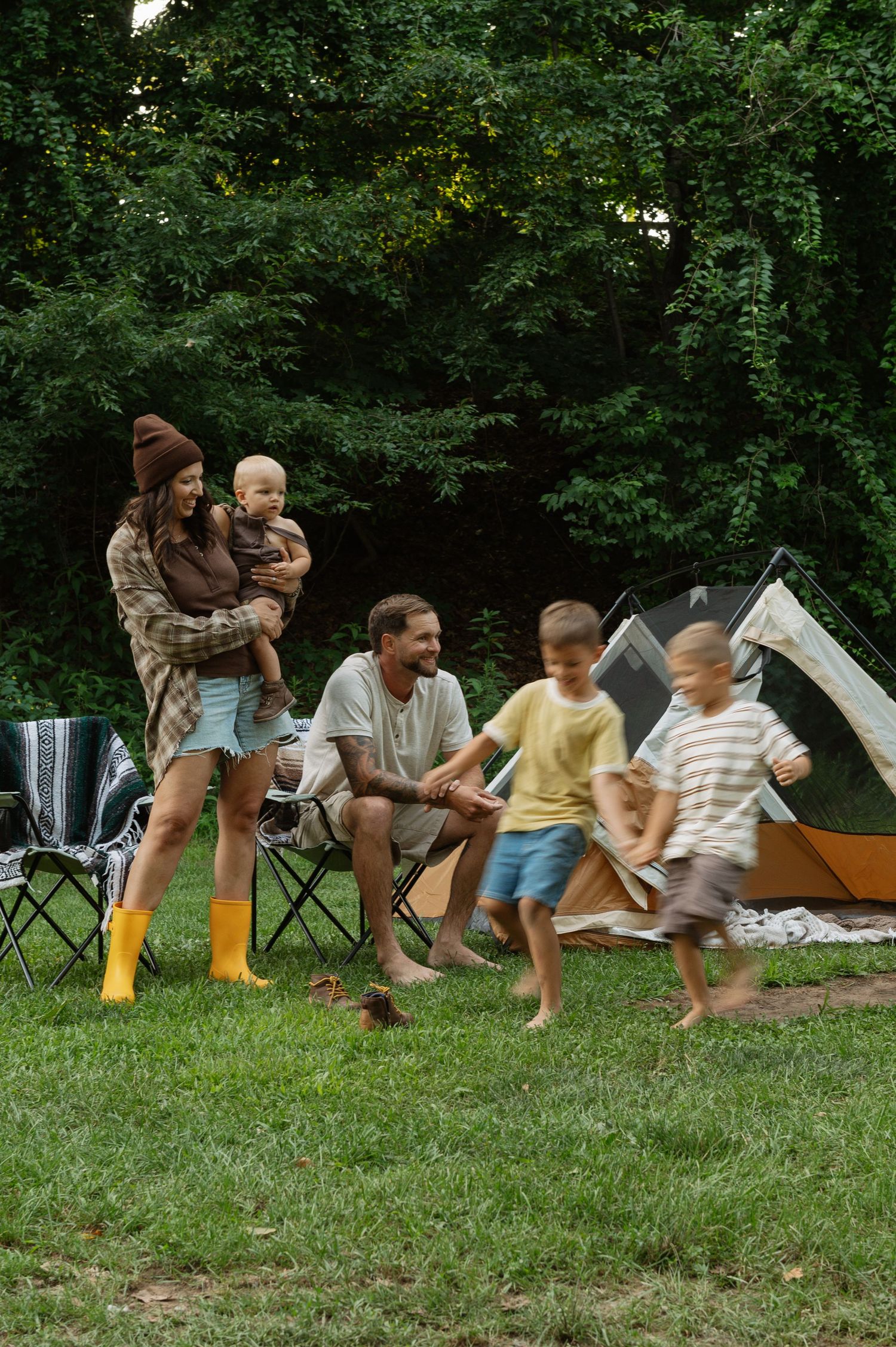 Family camping in forest with tent, parents watching children play on green grass near folding chairs.
