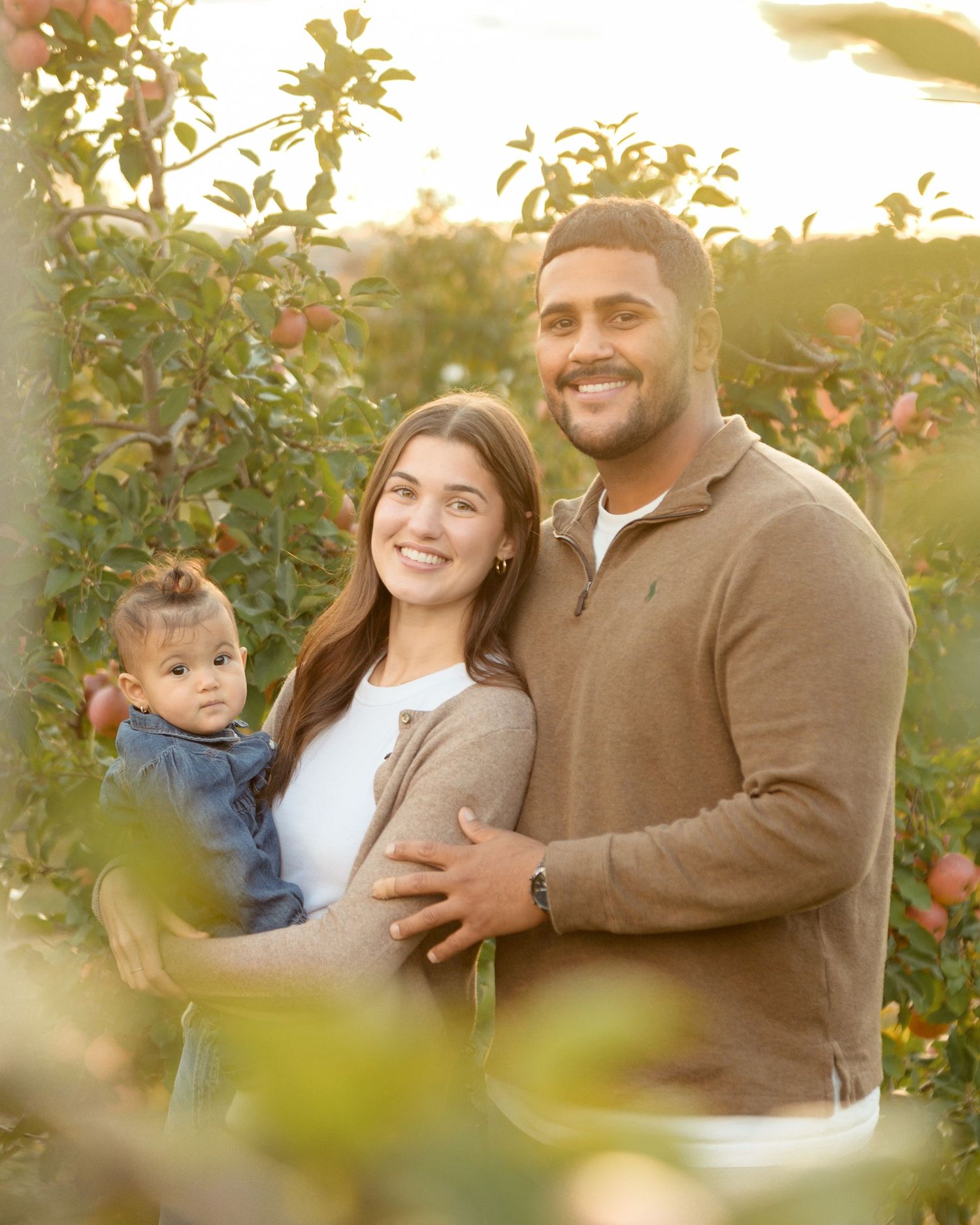 A couple and baby share tender moments among apple trees at sunset.