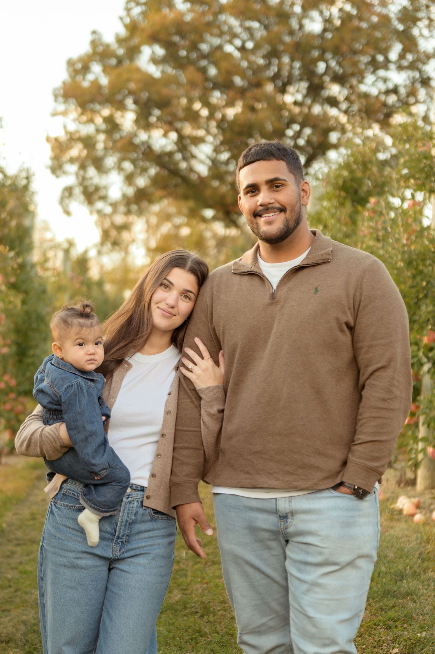 Family enjoying a warm autumn day together in a sunlit orchard setting wearing casual denim and brown attire.