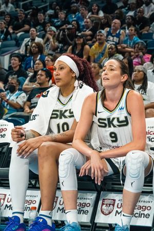 Professional basketball players in white jerseys sit on the sideline bench during a WNBA game.