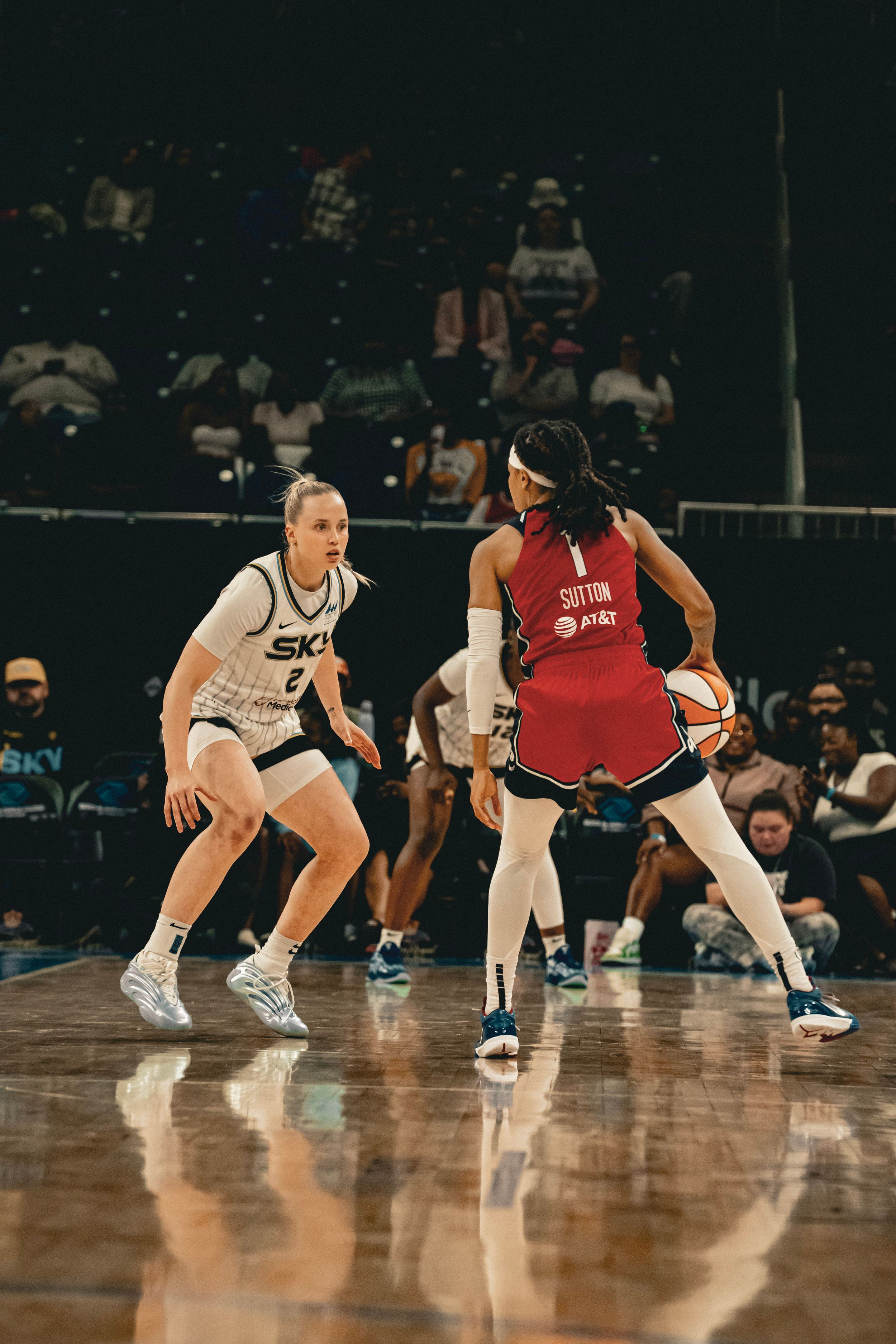 Two basketball players in a defensive stance during a WNBA game with one in white and one in red uniforms.