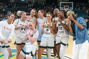Women's basketball team shares joyful moment together while celebrating in white uniforms on home court.