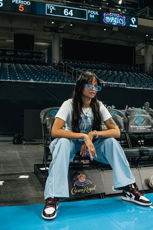Someone wearing sunglasses and a casual outfit sits in courtside seats at an empty arena with Crown Royal signage.