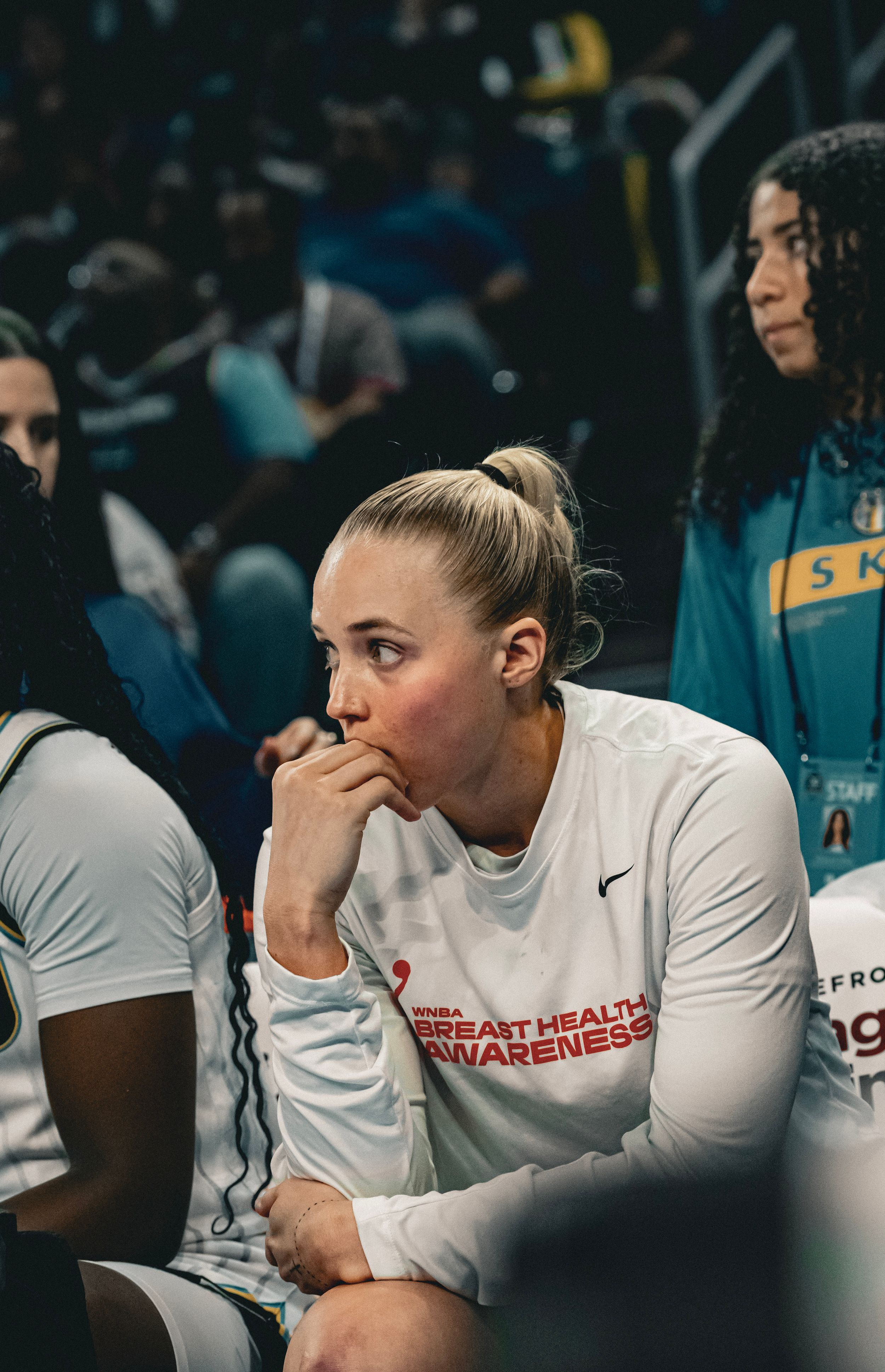 Basketball players in white Nike uniforms sit on the bench during a game.
