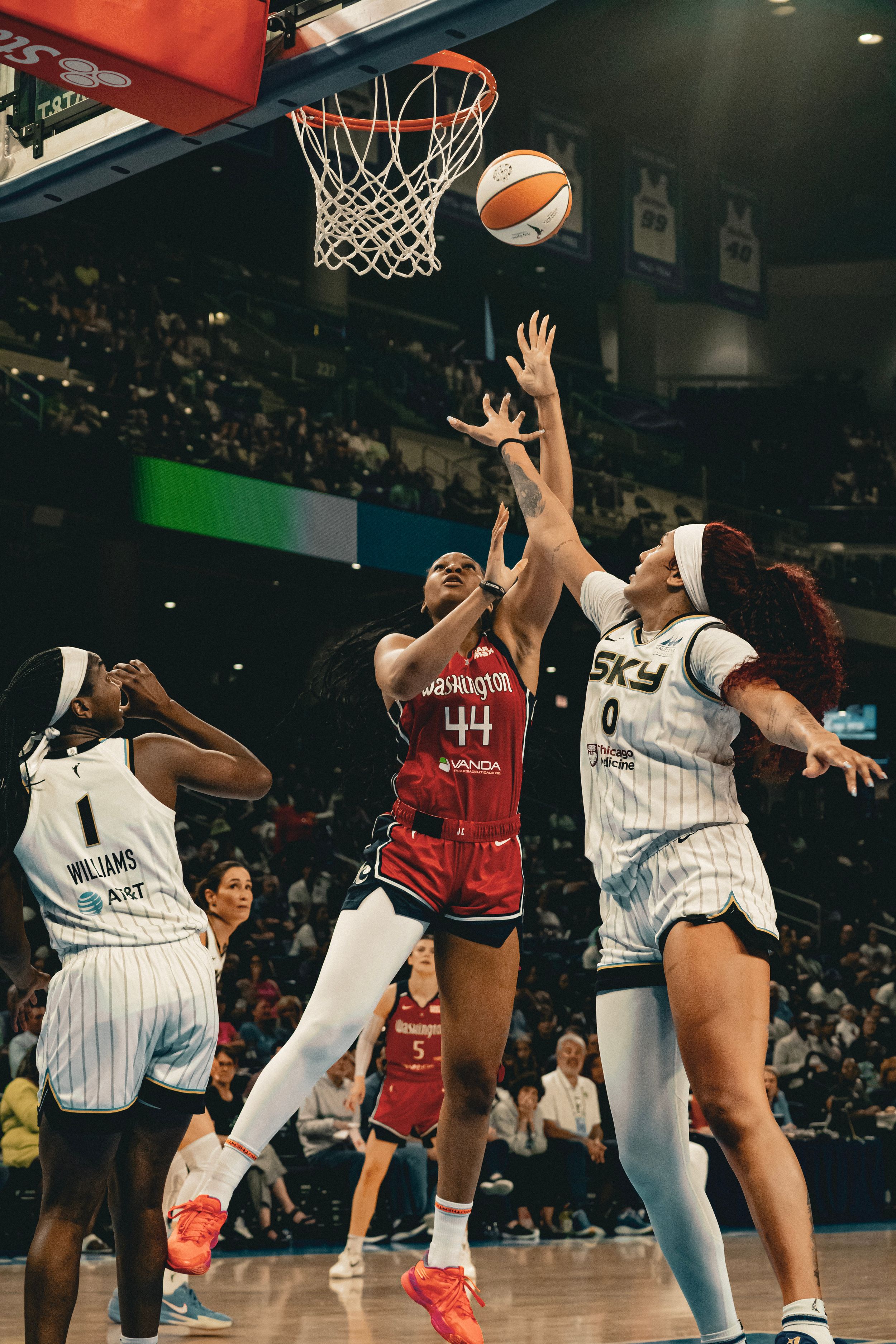 Basketball players leap for the ball during an intense WNBA game between opposing teams in white and red uniforms.