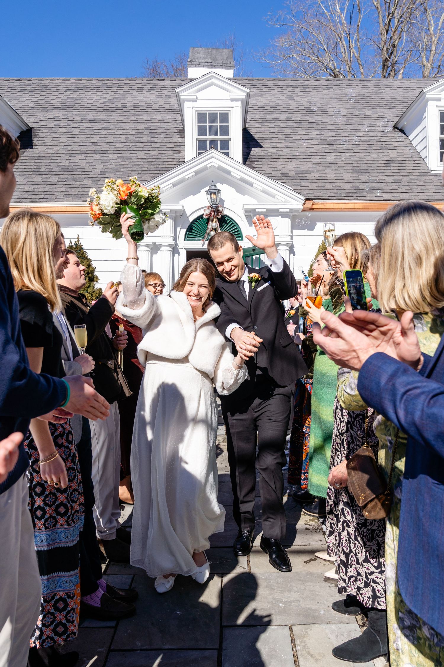 A group of wedding guests form a line outside a white house to celebrate and toast with champagne glasses.