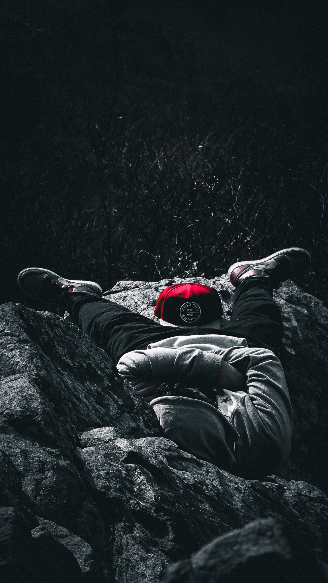 Person lying on dark rocky surface wearing black and red clothing in dramatic black and white lighting.