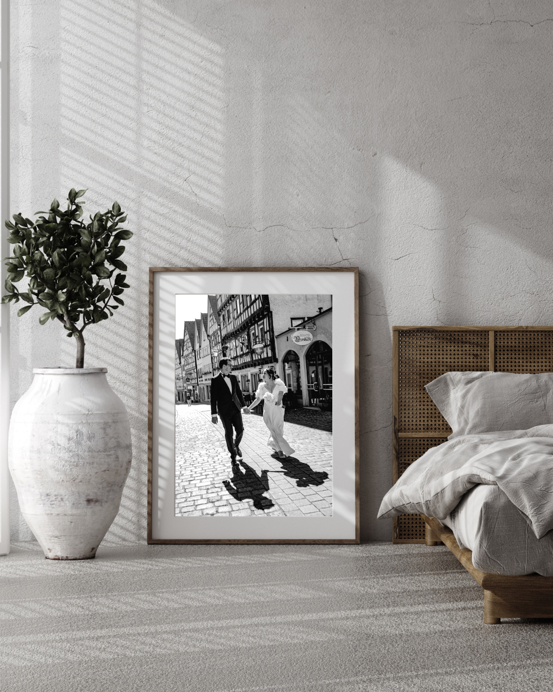 A minimalist bedroom scene with black and white framed artwork, a ceramic vase, and sunlight filtering through window blinds.