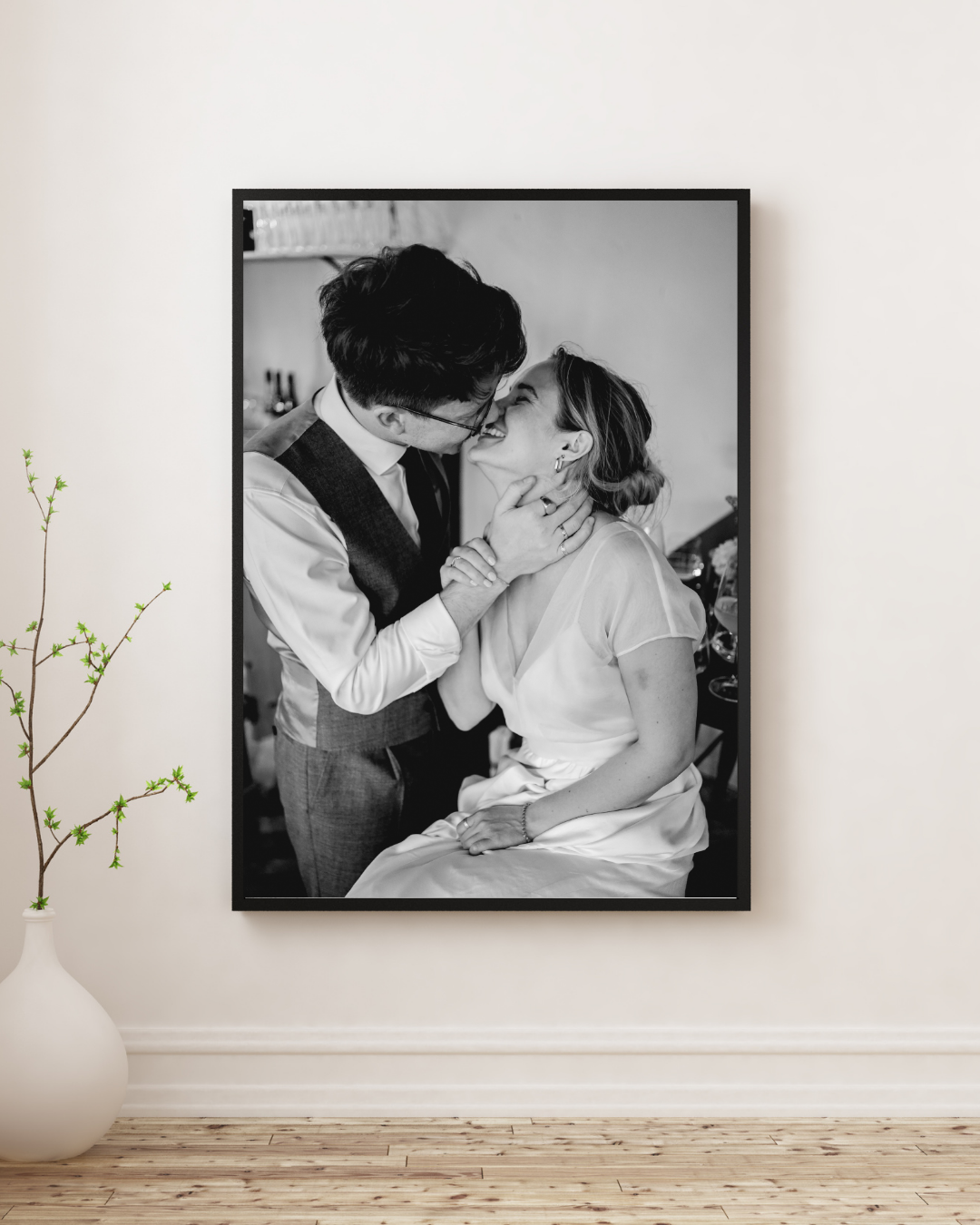 A white vase with delicate green branches sits beside a black and white framed photograph on a light colored wall.