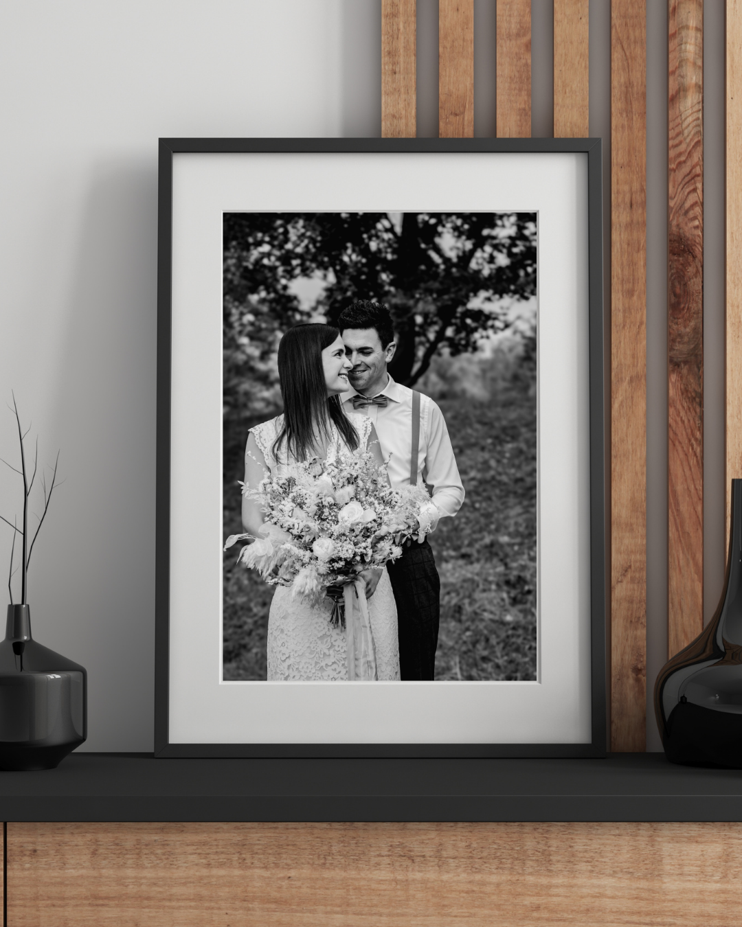 Display of framed black and white wedding photographs on wooden plank wall with decorative shelf.