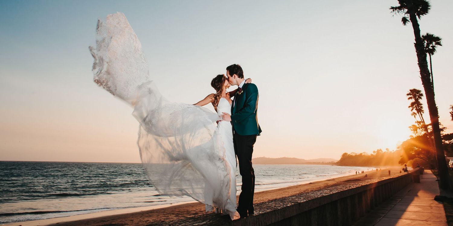 Couple kisses on beach at sunset as bride's dress billows dramatically in the wind in Santa Barbara at the Four Seasons Resort.