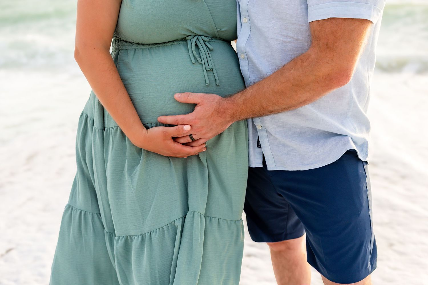 Couple tenderly holding pregnant belly together on white sand beach in matching green and blue outfits.