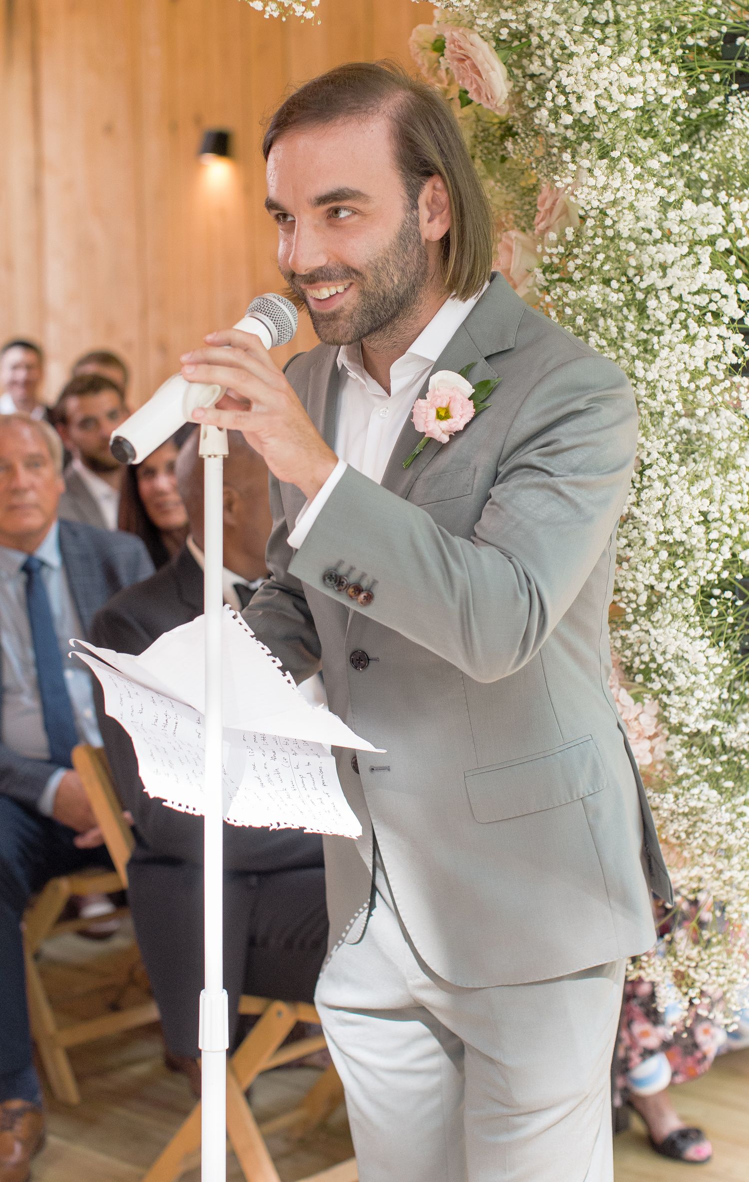 A person in a gray suit performs an energetic dance move during a celebration in a venue decorated with floral elements.