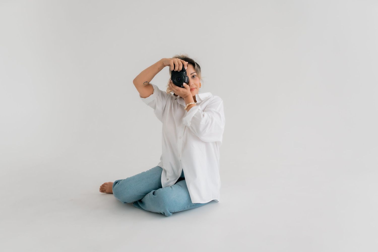 Photographer in white shirt and blue jeans sits cross-legged on white floor while taking a picture with vintage camera.