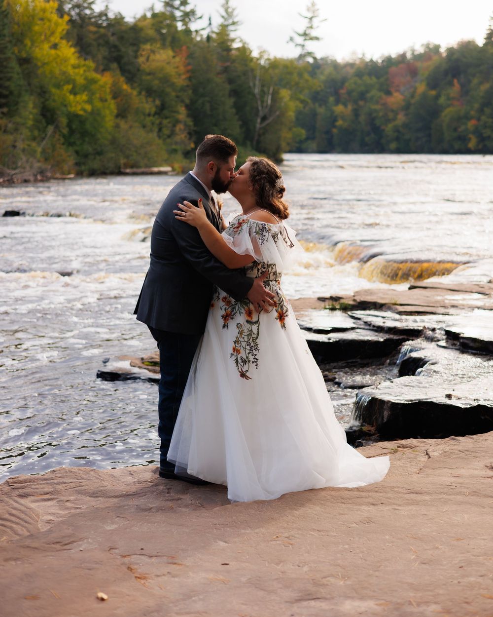 A couple shares an intimate moment on a rocky shoreline with a serene lake and autumn forest in the background.