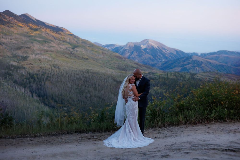 Bride and groom embrace on a mountain overlook at dusk with scenic Colorado peaks and forest in the background.