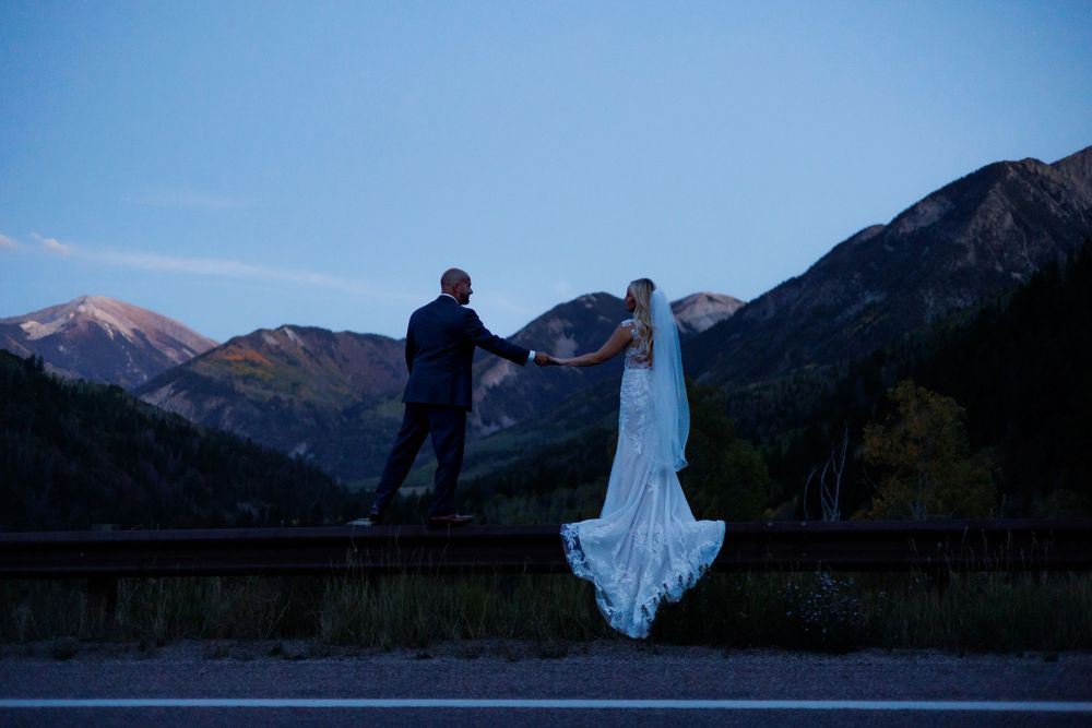 Bride and groom hold hands on a guardrail at dusk with stunning Colorado mountain peaks glowing in the background.