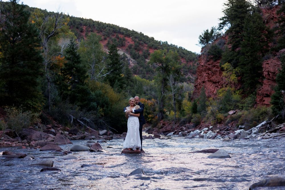 Couple embracing in white wedding attire on rocky riverbed surrounded by red canyon walls and autumn trees.