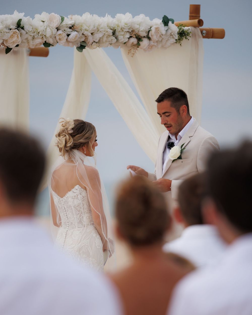 Couple exchanges vows under a floral arch during an outdoor wedding ceremony with guests in white attire.