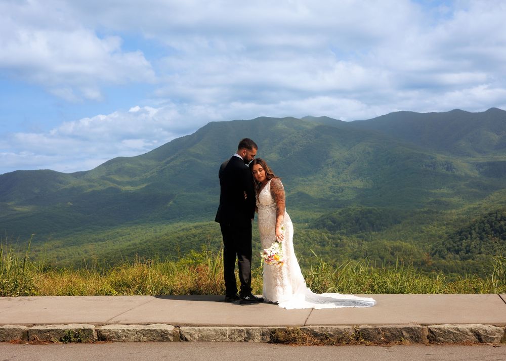 Bride and groom embrace at mountain overlook with lush green peaks and blue sky backdrop on their wedding day.