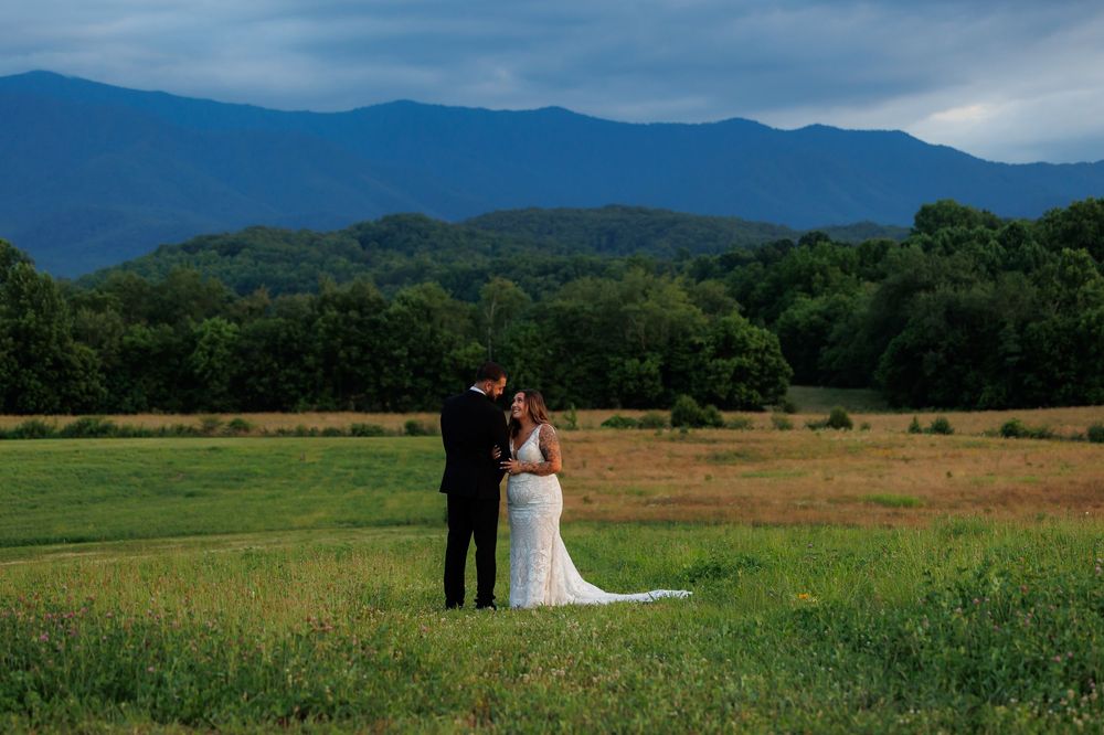 Bride and groom embrace in a lush green field with stunning Blue Ridge Mountain backdrop under dramatic stormy skies.