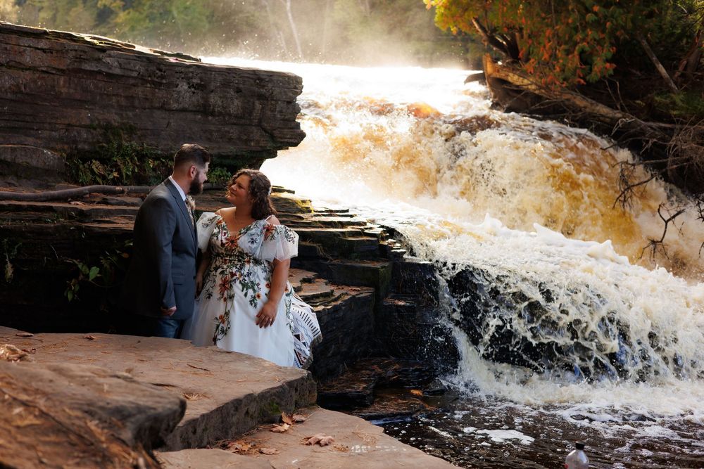 A couple in wedding attire poses together on stone steps near a cascading waterfall in a scenic outdoor setting.