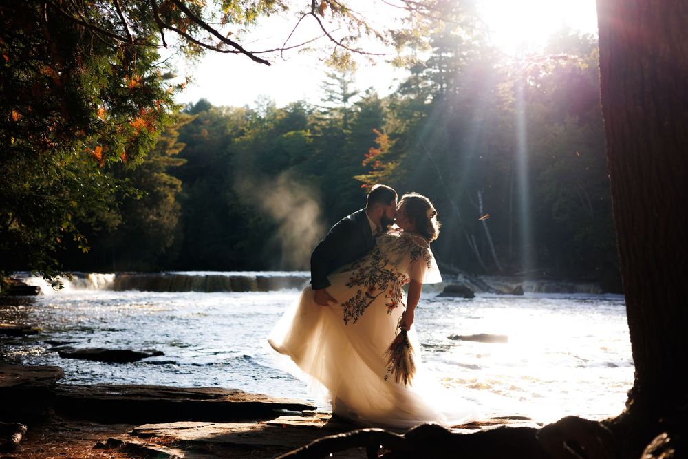 Romantic couple embraces at sunset by a serene lake surrounded by pine trees with sunlight streaming through branches.