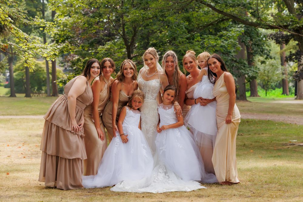 Bridesmaids in beige dresses gather outdoors with the bride in a white gown for wedding photos in a park setting.