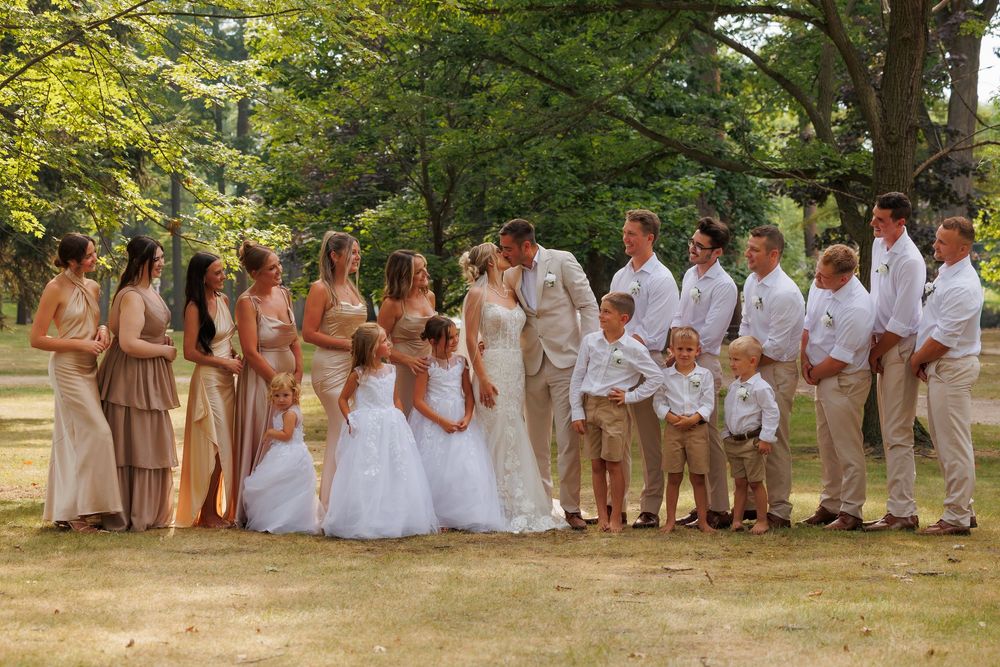 Wedding party in casual summer attire poses outdoors with bridesmaids in tan dresses and groomsmen in white shirts.
