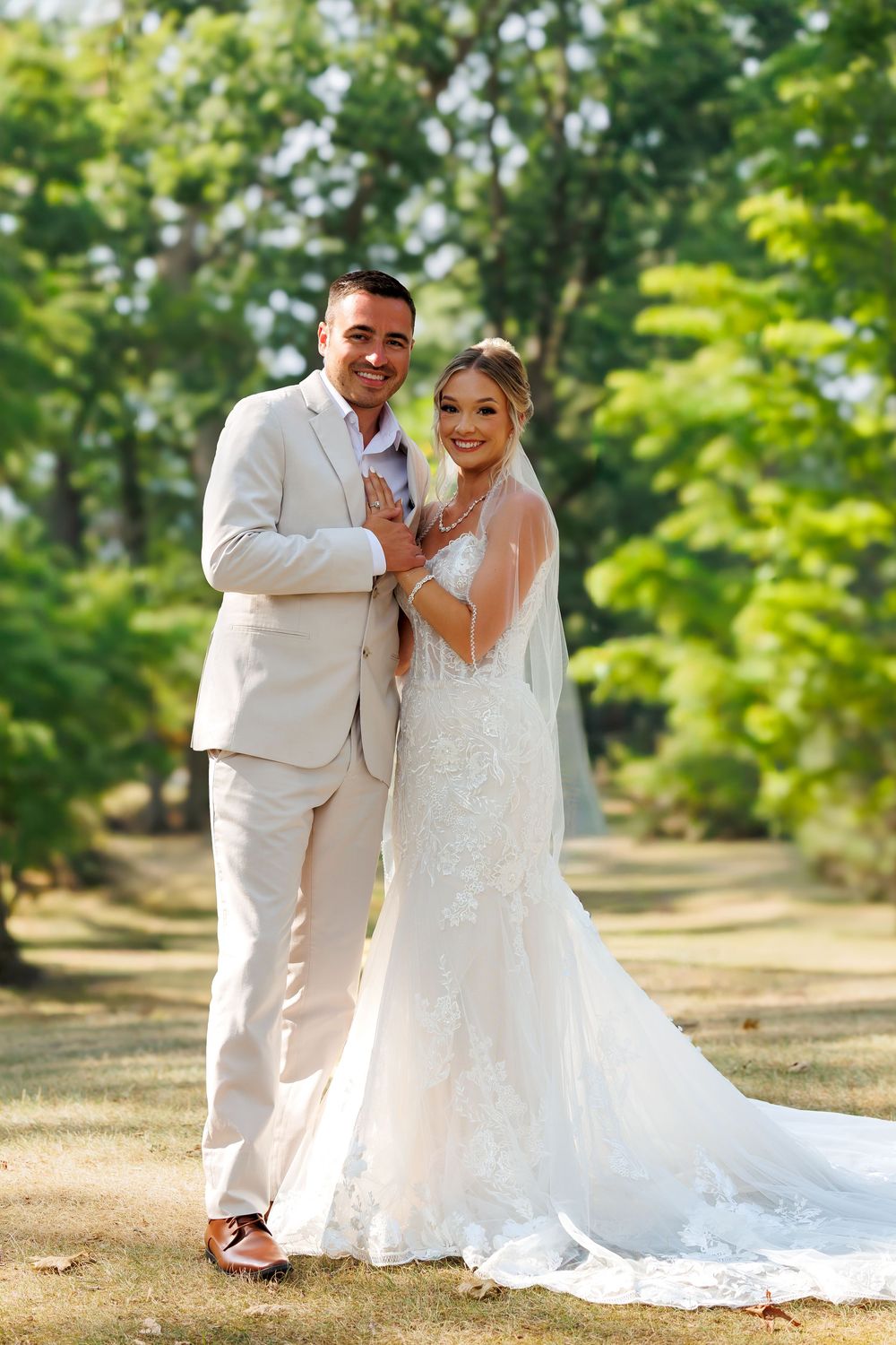 A wedding couple in elegant white attire poses together on a sunny day with lush greenery in the background.