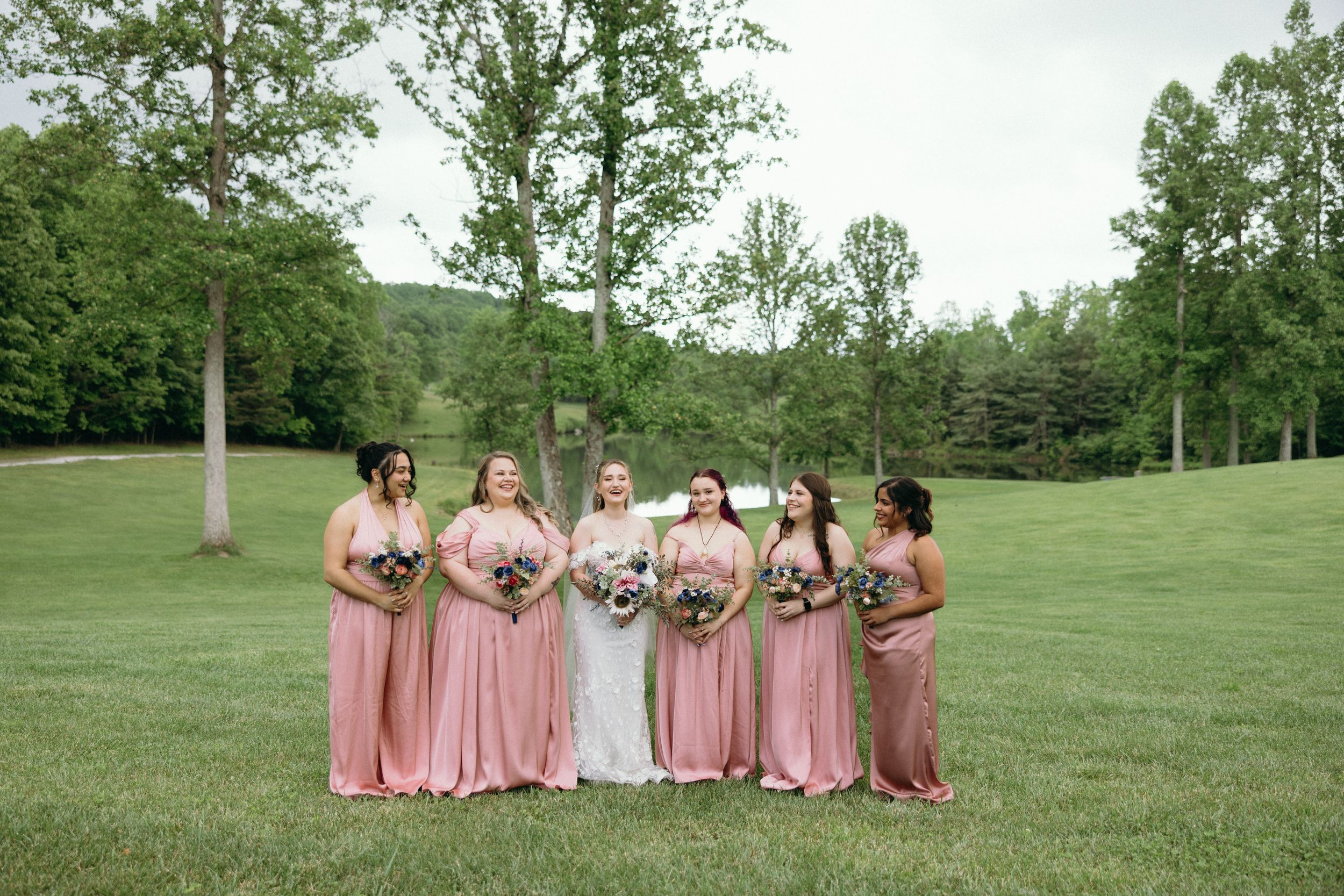 Wedding party in pink dresses poses together on a grassy hillside for group photos. Estate and historic venue wedding photographer for NC