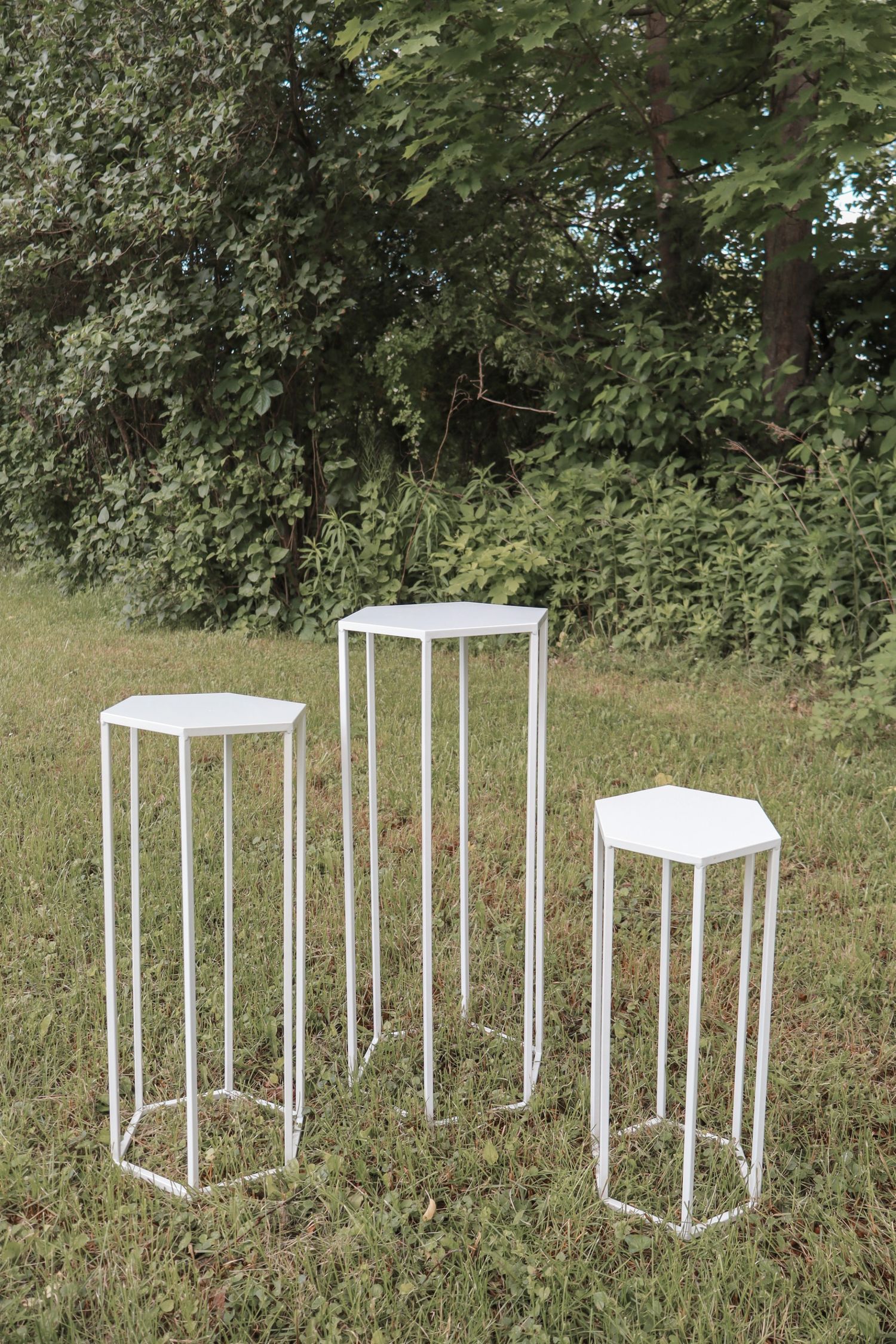 Three white metal geometric pedestals of varying heights stand on grass with trees in the background.