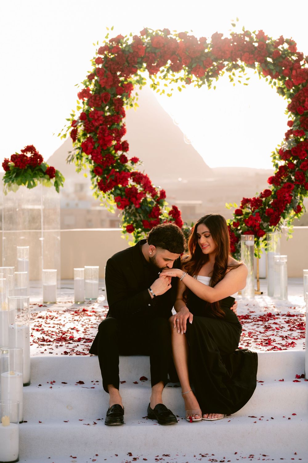 Romantic couple in black outfits poses near white columns with red rose petals scattered on the ground at sunset.