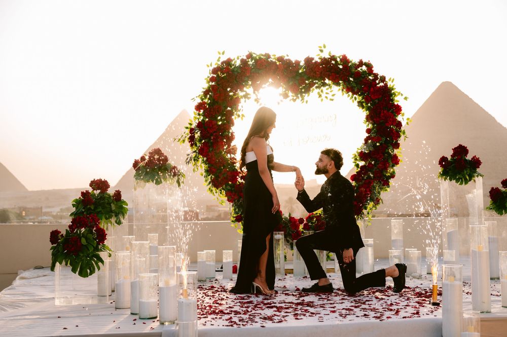 A row of white arbors decorated with red roses and greenery create a romantic outdoor wedding ceremony setup at sunset.
