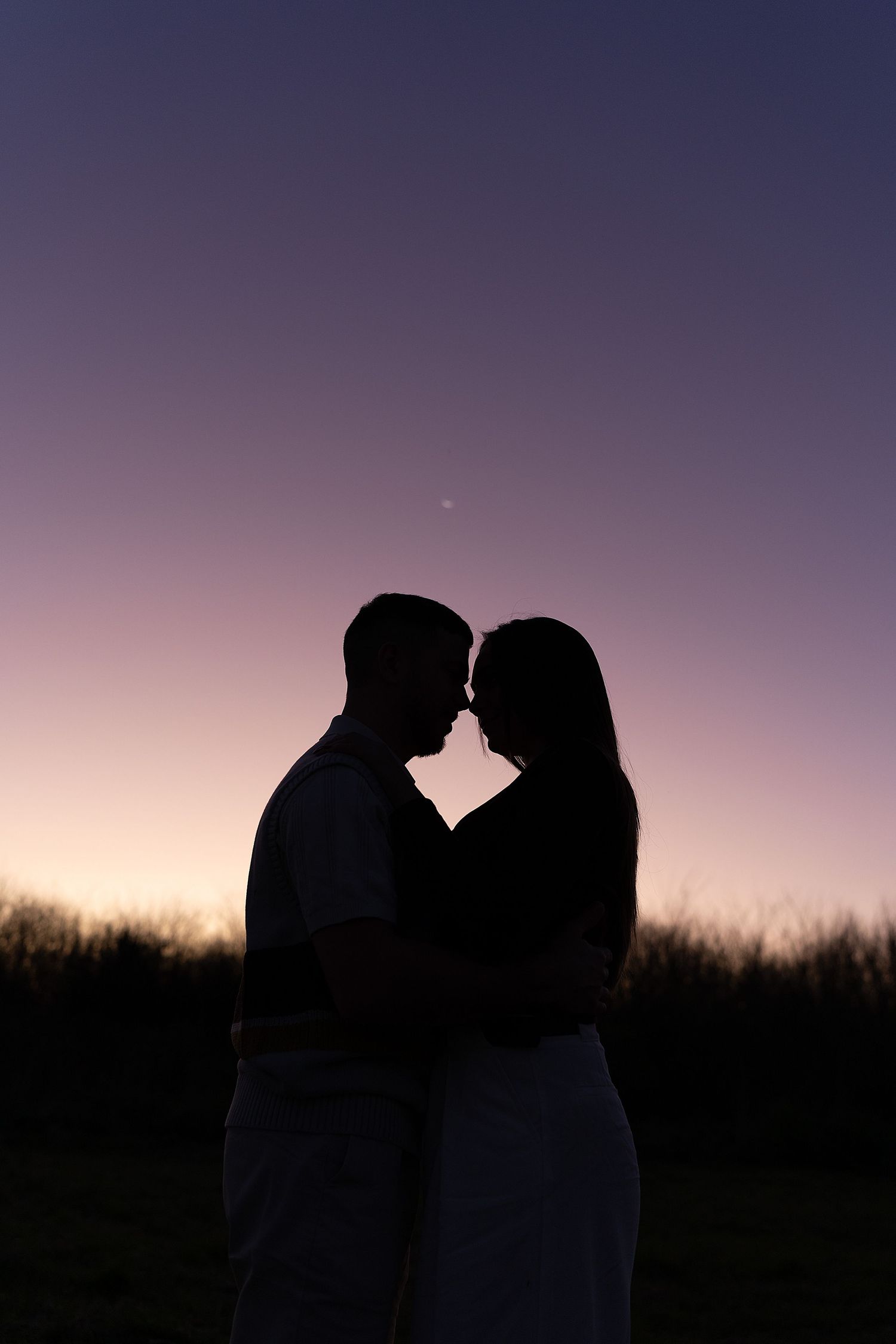 Silhouettes of a romantic couple kissing against a purple twilight sky beside a grassy field at sunset.