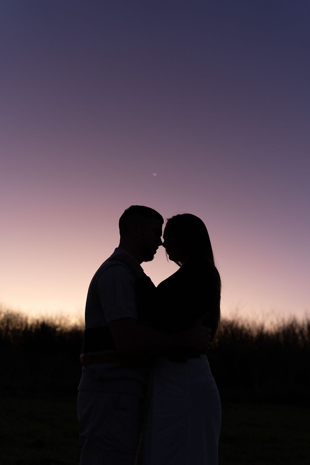 Silhouettes of a romantic couple kissing against a purple twilight sky beside a grassy field at sunset.