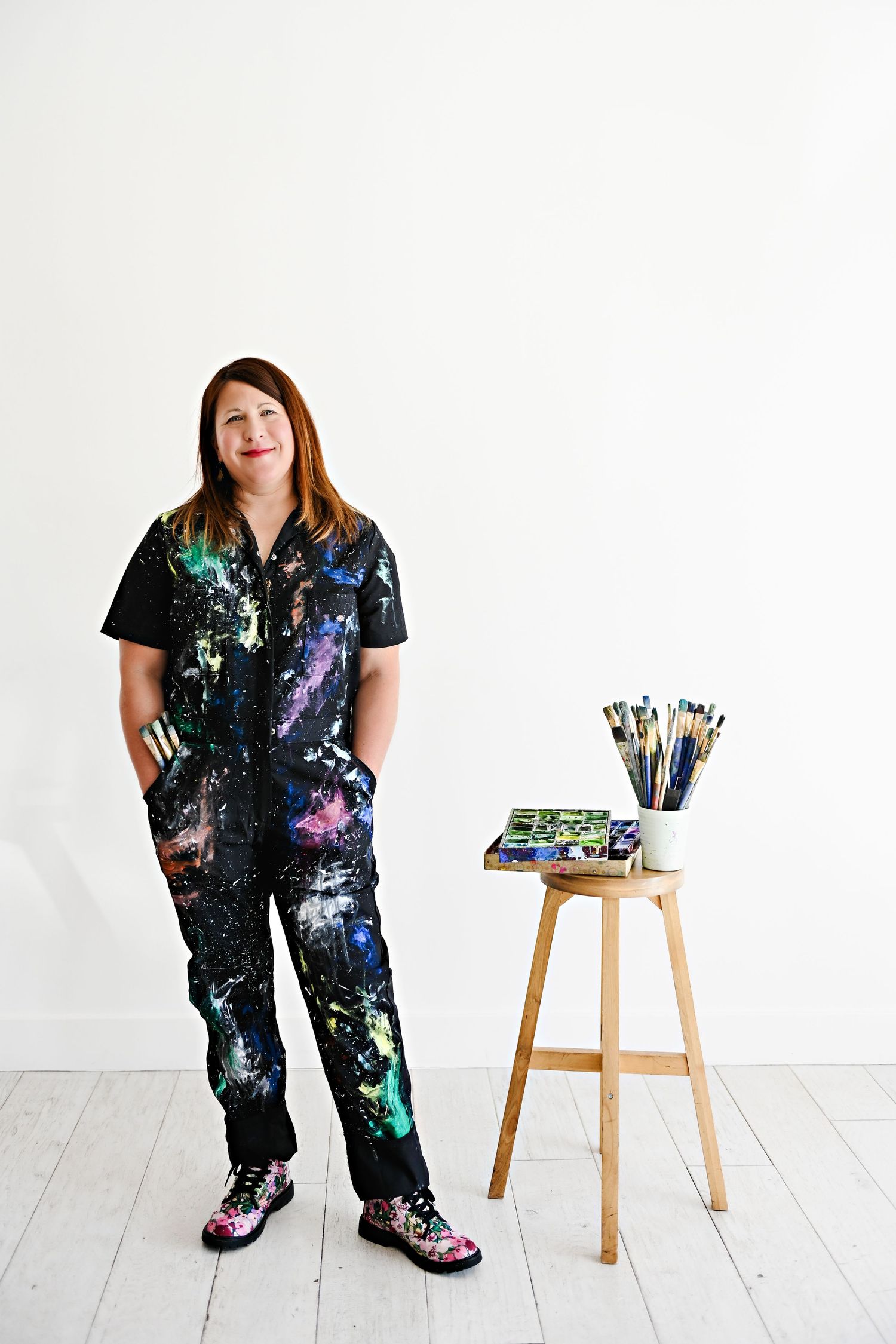 Artist wearing colorful paint-splattered jumpsuit posing with art supplies on wooden stool.
