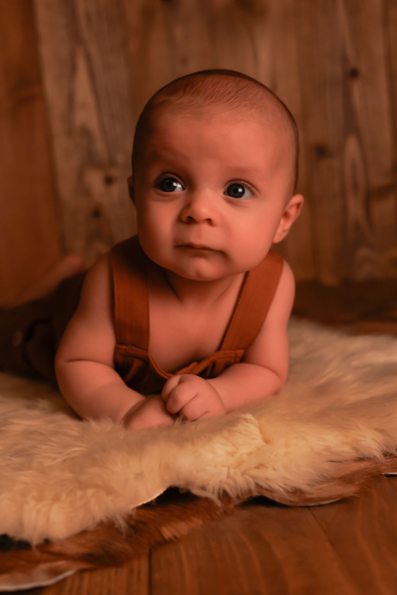 A baby in brown overalls lies on a white furry rug against wooden paneling.