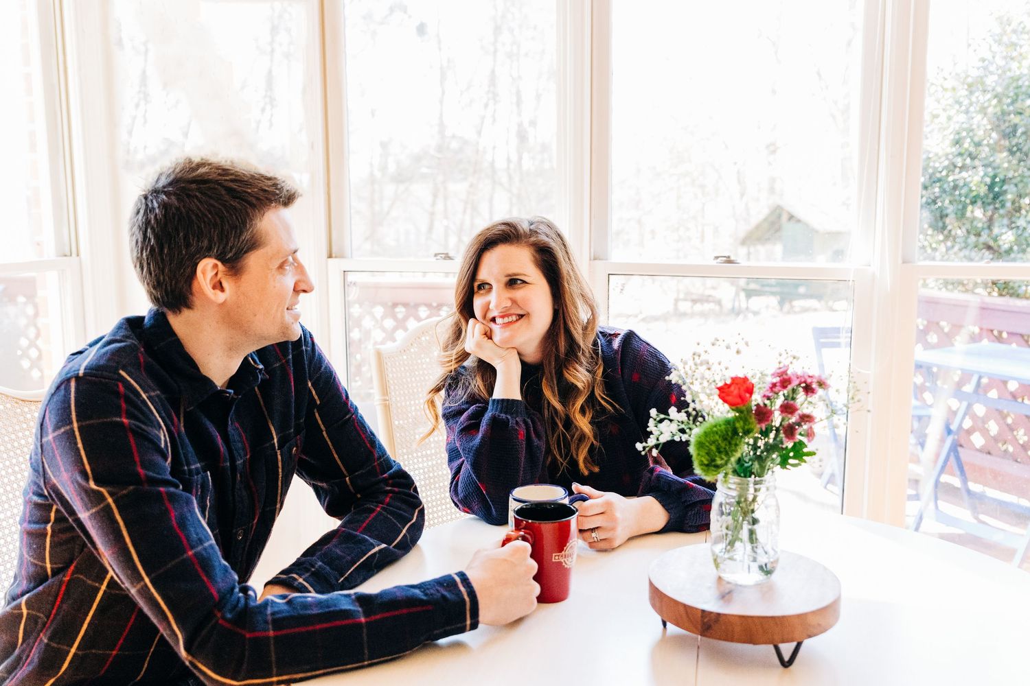Couple enjoying coffee together at sunny window table with red roses in vase creating romantic atmosphere.