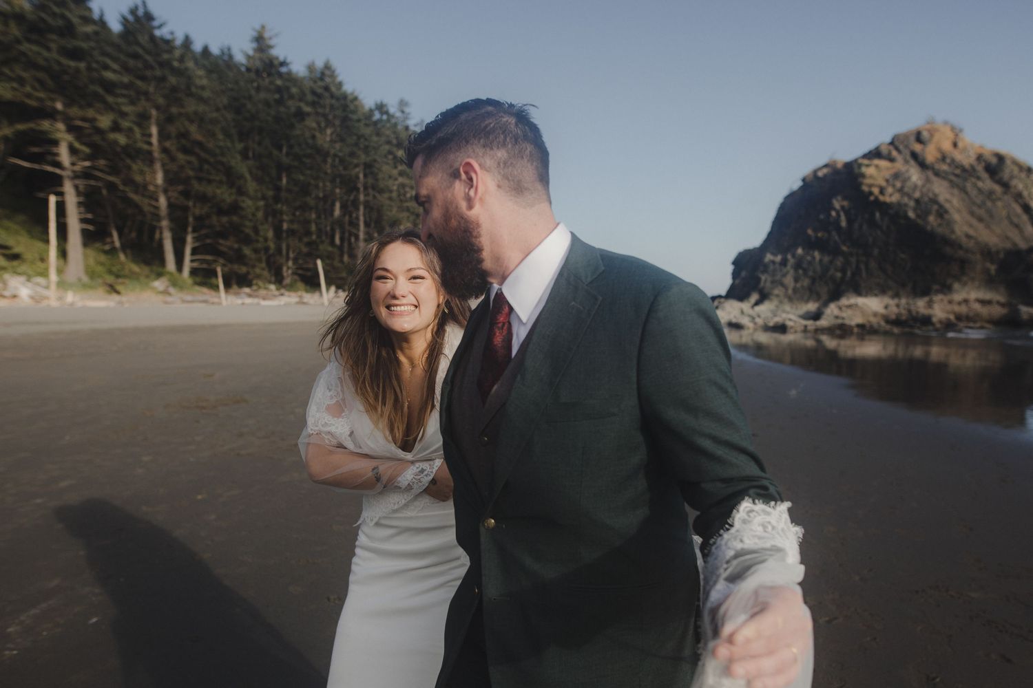 A couple shares a candid moment together on a scenic beach with rocky cliffs and evergreen trees in the background.
