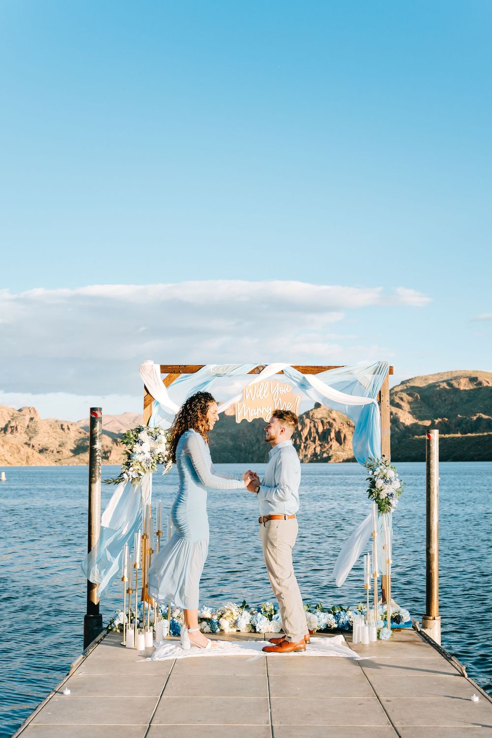 A couple stands under a decorated wedding arch on a dock with mountains and lake in the background.