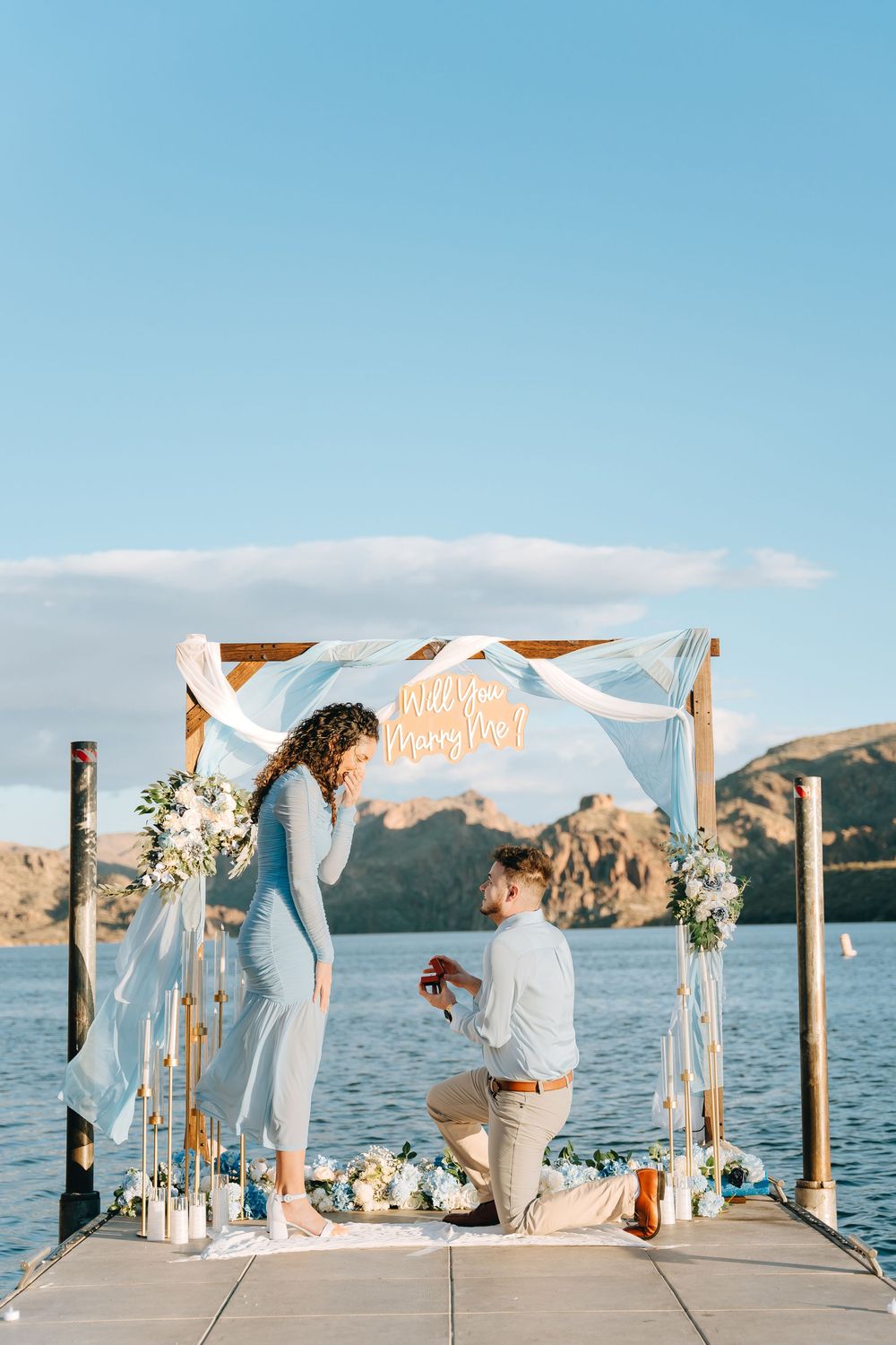 A proposal scene on a lakeside dock with a wooden arch decorated with white fabric and flowers against mountain views.