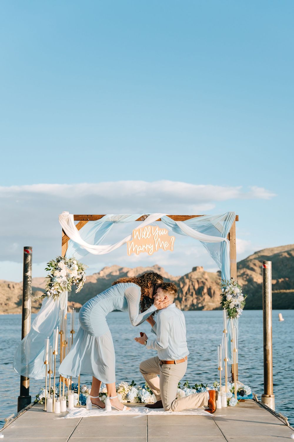 A couple shares an intimate moment during a lakeside proposal under a decorated wooden arch.