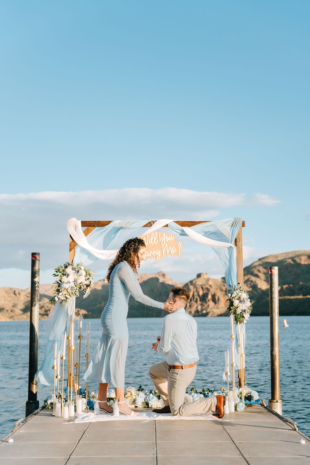 A romantic proposal moment unfolds on a decorated dock overlooking a scenic mountain lake.