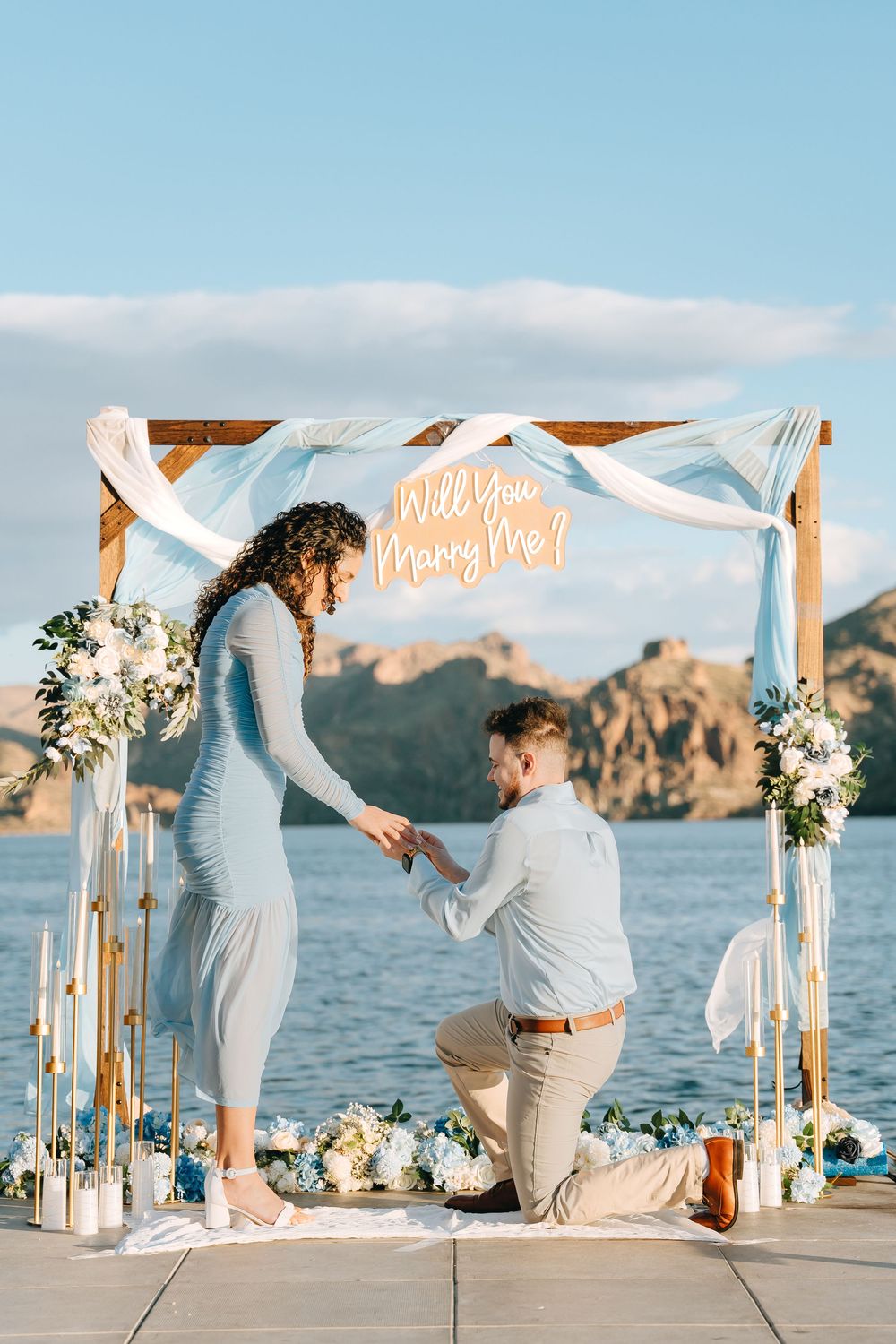 A heartfelt proposal takes place on a dock with mountains and lake views under a decorated arch.