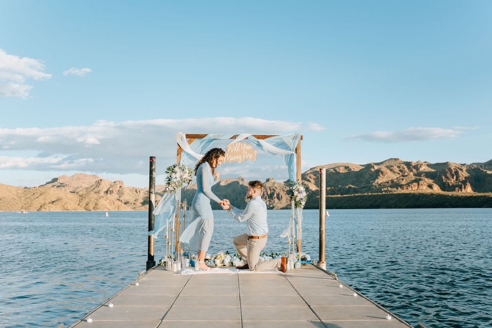A couple celebrates their engagement on a scenic dock with mountains and lake in the background.