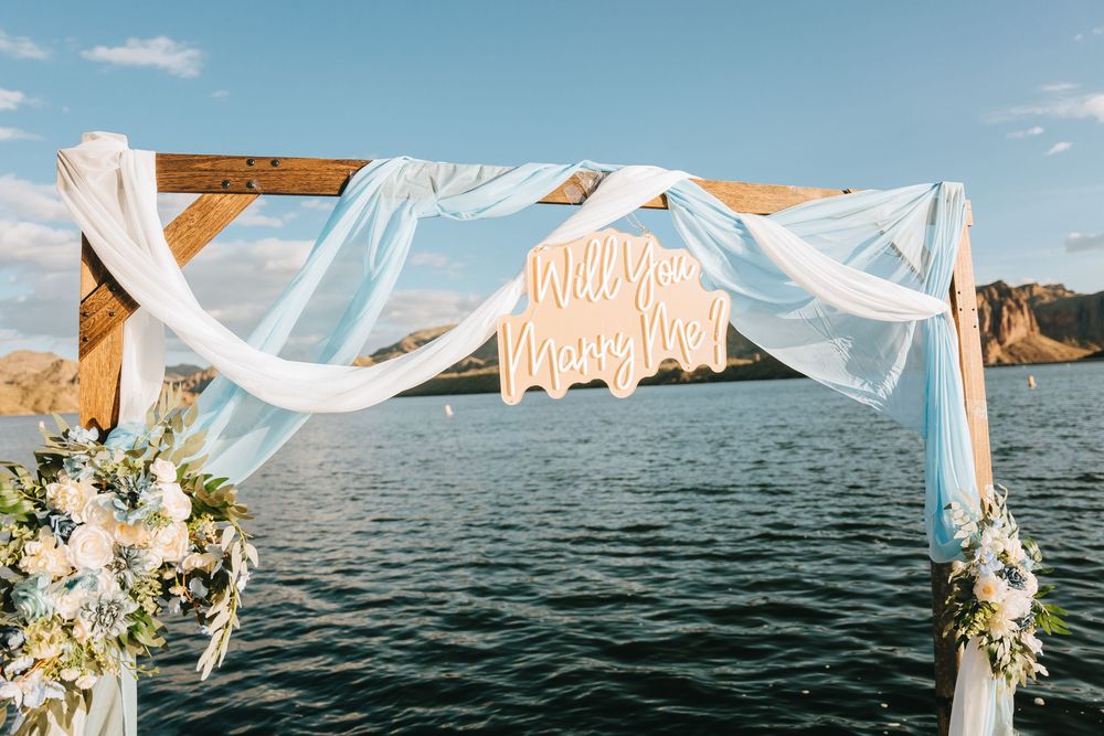 A wooden wedding arch decorated with white fabric and floral arrangements overlooking a scenic lake.