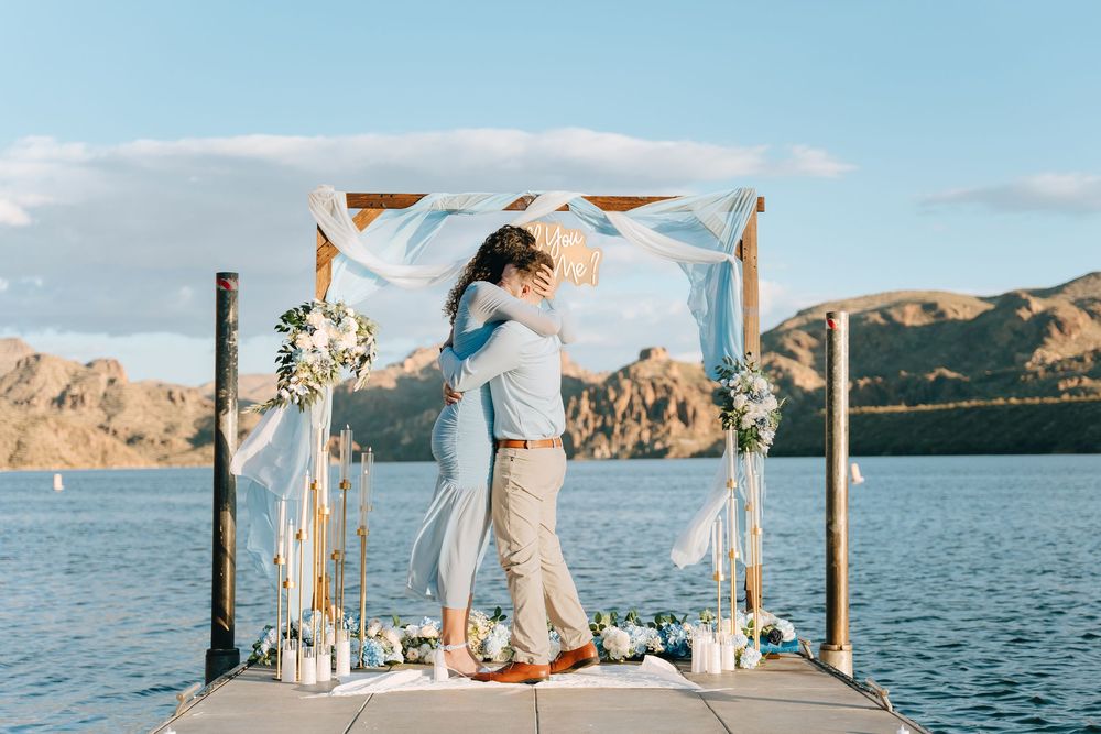 Two people embrace joyfully under an arch decorated with flowing fabric and flowers by the lake.
